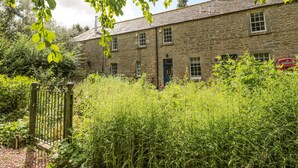 A stone building with multiple windows and a blue door, surrounded by dense greenery and a metal gate. - The Coach House (Duns)