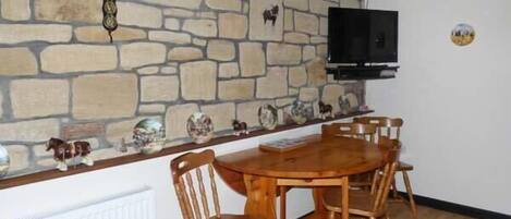 A dining area with a stone wall, wooden ceiling beams, a TV mounted above the wall, and a table with chairs.