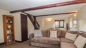 A living room with a brown sectional sofa, a wooden bookshelf, and a staircase. - Woodside Barn (Ulverston)