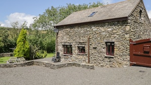 A stone building with a sloped roof, two windows with red frames, a satellite dish, and a gravel driveway. - Woodside Barn (Ulverston)