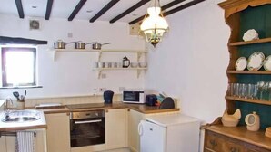 A compact kitchen with a sink, stove, microwave, and a small refrigerator. There is a wooden table with a bowl and chairs. - The Cottage (Buxton)