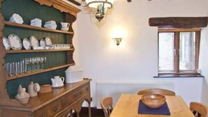 A traditional dining room with a wooden table, chairs, and a wall-mounted shelf with dishes and glasses. - The Cottage (Buxton)