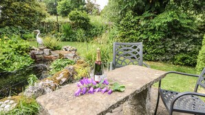 Un tavolo in pietra con una bottiglia di champagne e fiori viola, circondato da un giardino con uno stagno, rocce e vegetazione.