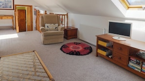 Une pièce chaleureuse avec un plafond haut, un fauteuil confortable, une table d’appoint en bois, une bibliothèque, un téléviseur et une lucarne.