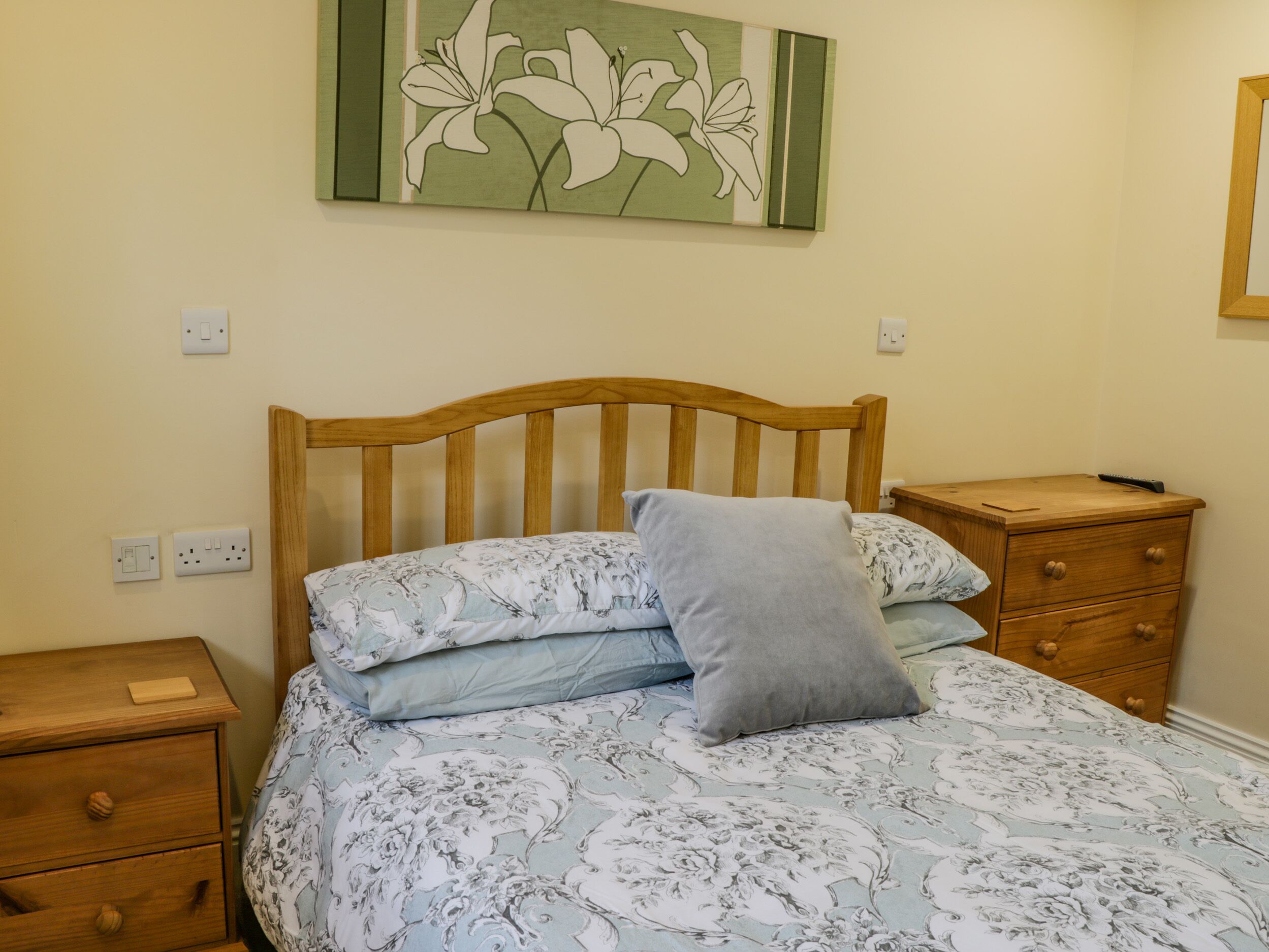 Une chambre à coucher avec une tête de lit en bois, une couvre-lit à motifs floraux, un oreiller gris, une table de chevet en bois avec une télécommande, et une commode en bois avec des tiroirs.