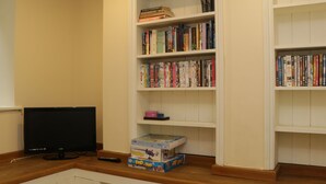 A room with a television on a wooden table, a bookshelf filled with books and DVDs, and a small box of games. - Apartment 6 (Whitby)