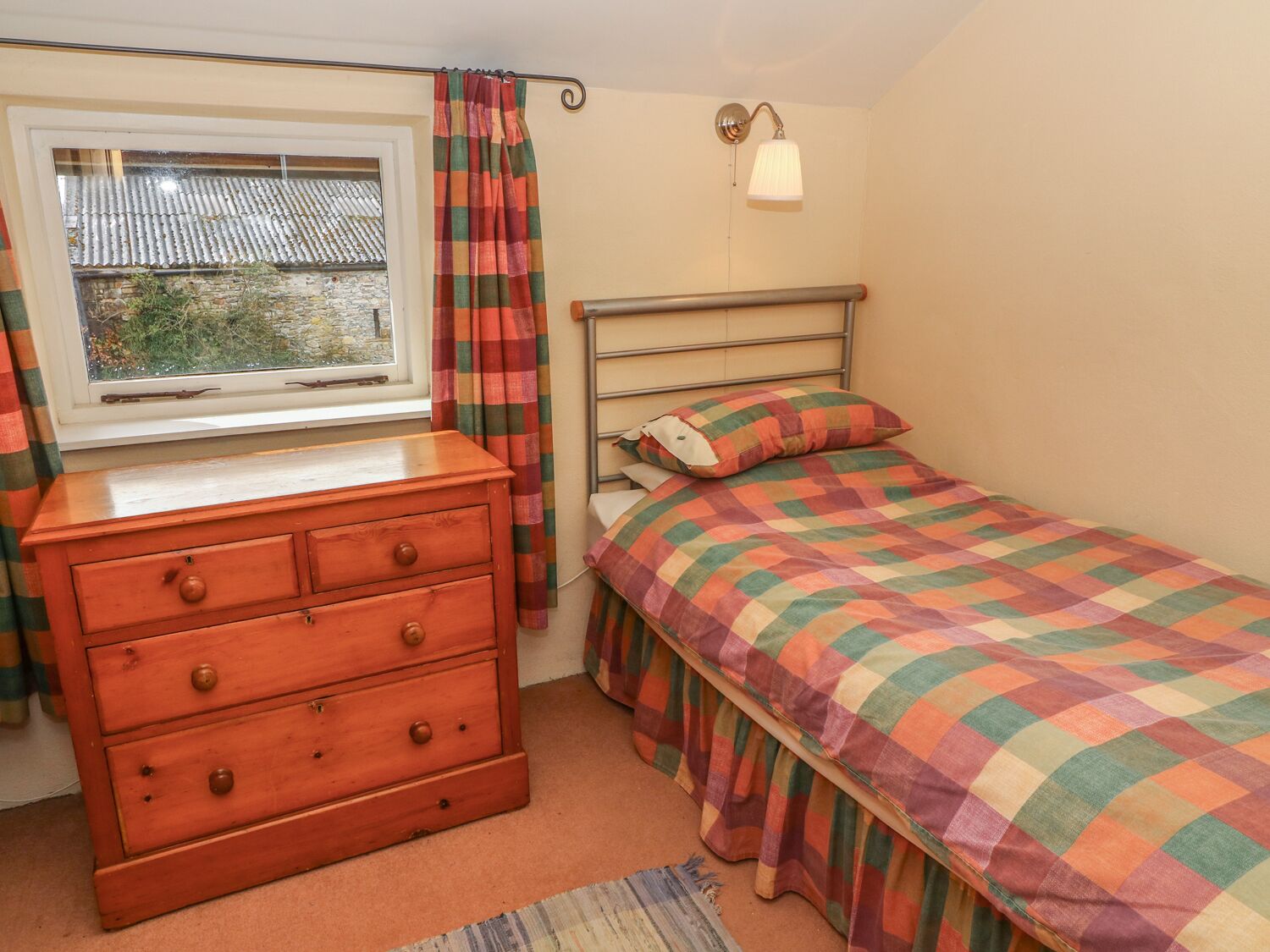 Un dormitorio con un aparador de madera, una cama con colcha a cuadros, una ventana con cortinas y una lámpara de pared.