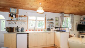 A compact kitchen with wooden cabinets, a dishwasher, a stove, and a dining area with a table and chairs. - The Log Cabin (Craven Arms)