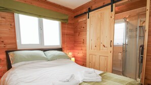 A wooden cabin room with a bed, a window with a green curtain, a wooden door, and a glass shower enclosure. - The Log Cabin (Craven Arms)