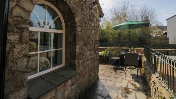 A stone building with a large arched window, outdoor patio with a table and chairs, and a green umbrella. - The Old Mill (Grange-over-Sands)
