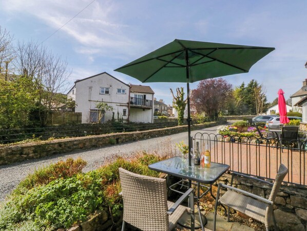 An outdoor patio with a table and chairs, a green umbrella, and a gravel path leading to houses. - The Old Mill (Grange-over-Sands)