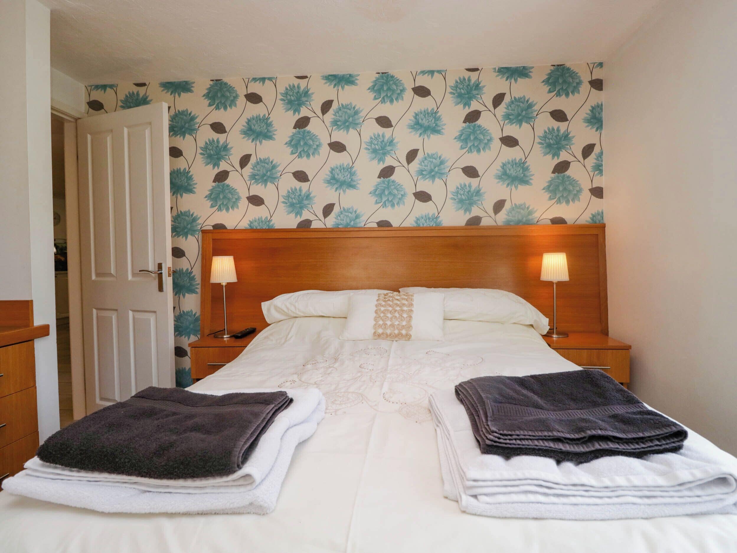 Een hotelkamer met een houten hoofdeinde, twee nachtkastjes met lampen, een bed met witte lakens en bloemenbehang.