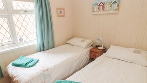 A bedroom with two single beds, a nightstand with a lamp and alarm clock, a window with curtains, and a painting of beach huts on the wall. - Sunnyside Cottage (Filey)