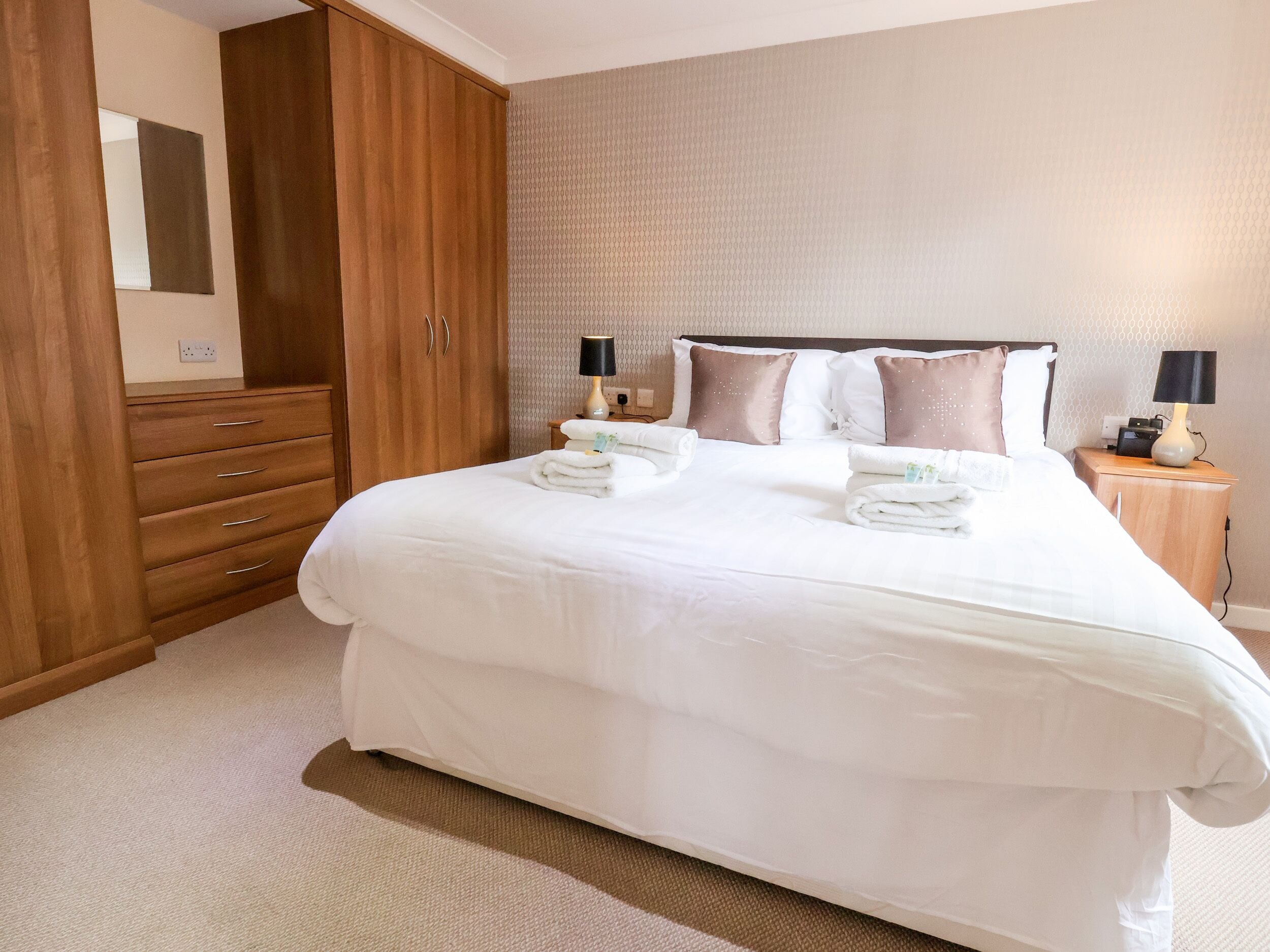 A neatly made bed with white linens, a wooden wardrobe, a bedside table with a lamp, and a patterned wall.