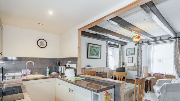 A modern kitchen with granite countertops, white cabinetry, and a built-in oven. There is a dining area with a wooden table and chairs, and a living room with a red rug and a white sofa. - Duck Cottage (Bridlington)