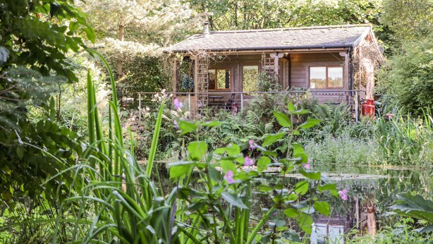 A rustic cabin with a wooden deck, surrounded by lush greenery and a pond. - The Log Cabin (Oban)