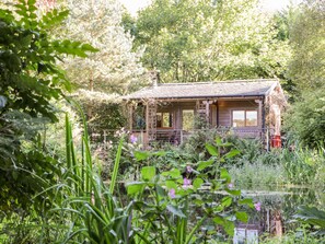 Un chalet rustique doté d’une terrasse en bois, entouré d’une végétation luxuriante et bordé d’un étang.