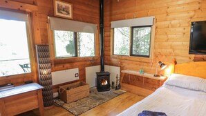A cozy cabin room with a wooden interior, a bed with a folded towel, a desk with a chair, a fireplace, and large windows. - The Log Cabin (Oban)