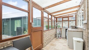 A sunroom with large windows, a wooden ceiling, a small kitchen area, and a seating area with a bench. - Violet Cottage (Thetford)