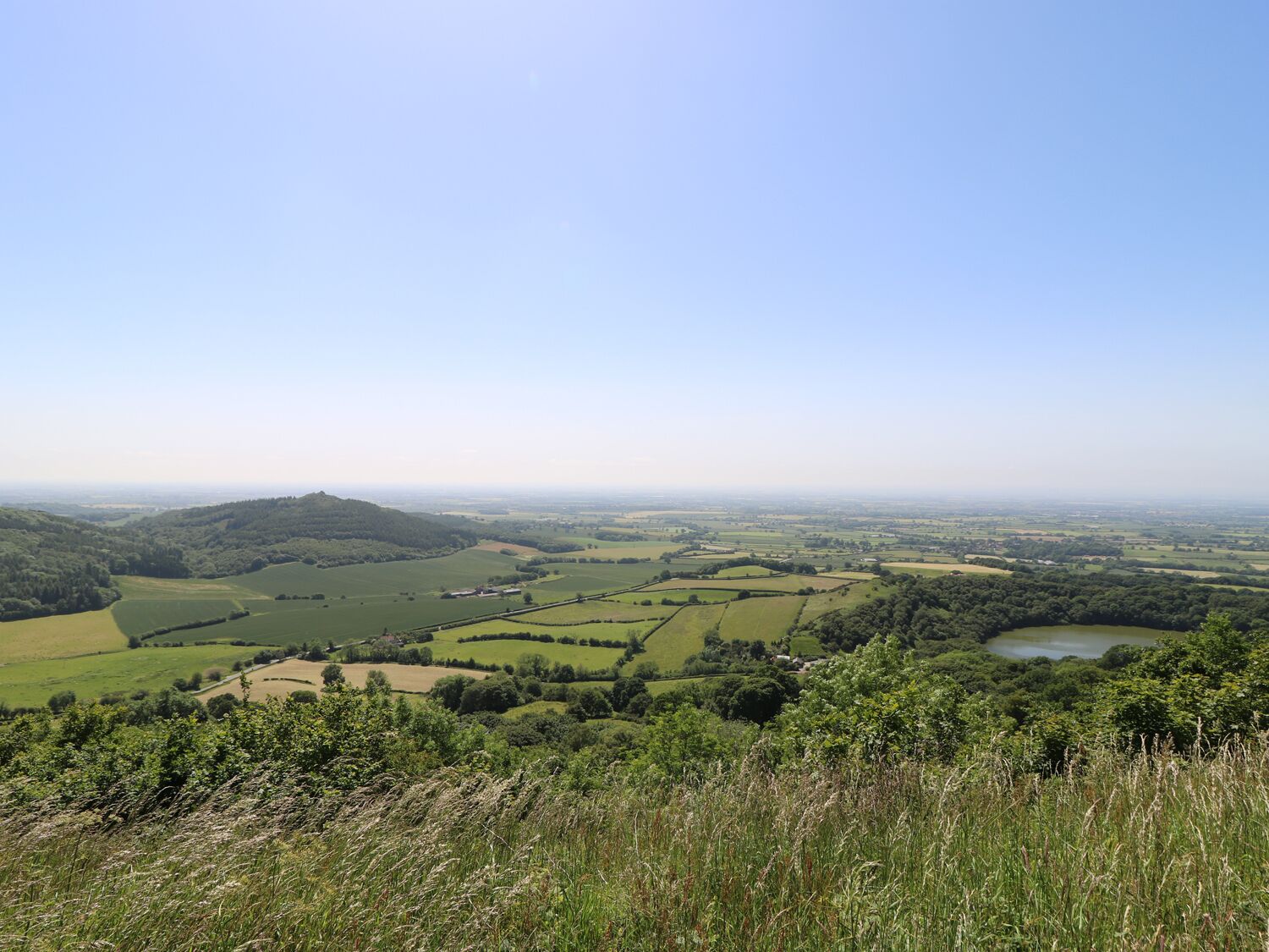 Une vue panoramique d’un paysage rural, avec des collines ondulantes, des champs et un petit plan d’eau.