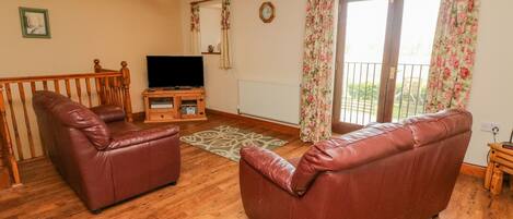 A living room with a wooden floor, two leather sofas, a TV stand, and a balcony with floral curtains.