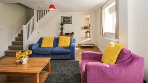A living room with a blue sofa, a purple armchair, and a wooden coffee table. There is a staircase with a white railing and a red pendant light. A kitchen is visible in the background.