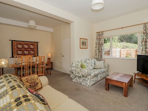 A living room with a sofa, a wooden coffee table, a dining table with chairs, a framed picture on the wall, and a window with curtains.