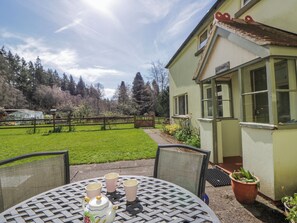 A patio with a table set for tea, a greenhouse in the background, and a house with a garden.