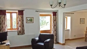 A spacious living room with a black leather sofa, a wooden cabinet, and a television. There are two windows with curtains, a radiator, and a chandelier. - The Stables (Hope Valley)