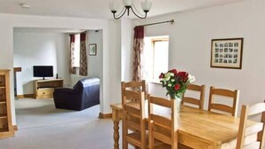 A dining area with a wooden table and chairs, a vase with flowers, and a television in the background. - The Stables (Hope Valley)