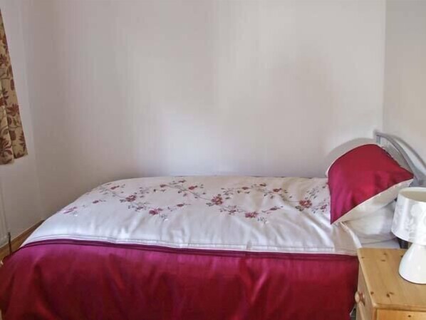 A bed with a floral bedspread and a red pillow, a bedside table with a white lamp, and a wooden nightstand. - The Stables (Hope Valley)