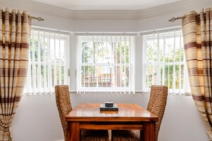 Comedor con mesa de madera, sillas de mimbre, ventanas amplias con persianas blancas y vista a zonas verdes.