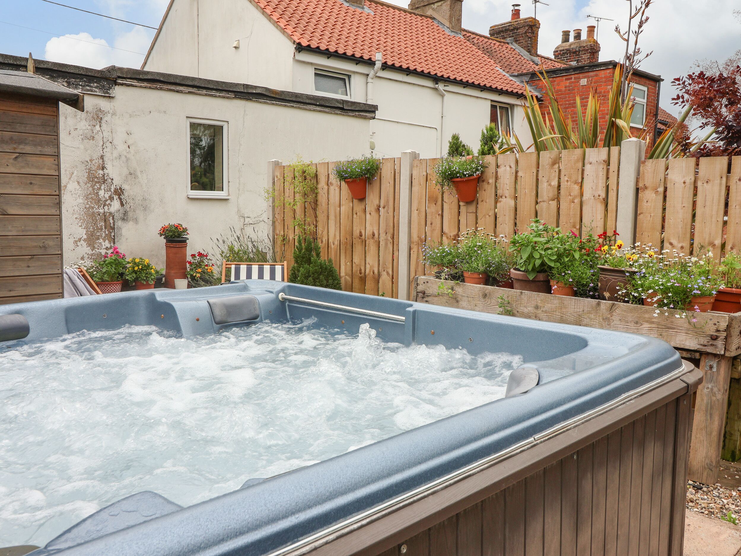 Un jacuzzi en un patio trasero con cerco de madera, plantas en macetas y una casa al fondo.