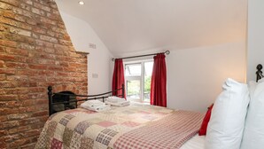 A bedroom with a brick wall, a quilted bedspread, and a window with red curtains. - The Cottage (Driffield)