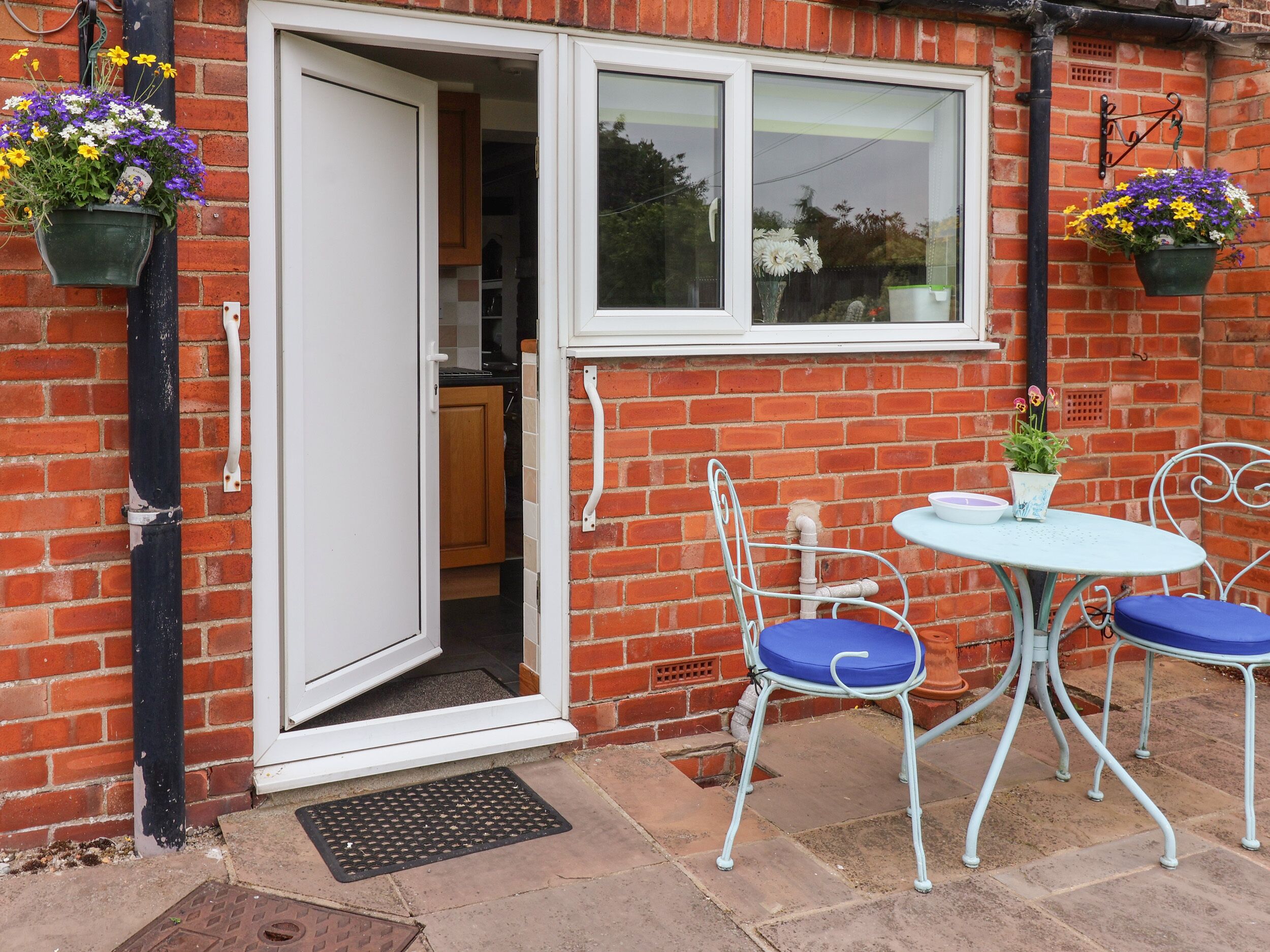 A brick house with a white door and window, a small patio with a round table and two chairs, and hanging flower baskets.