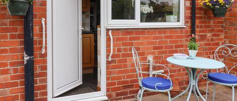 A brick house with a white door and window, a small patio with a round table and two chairs, and hanging flower baskets.