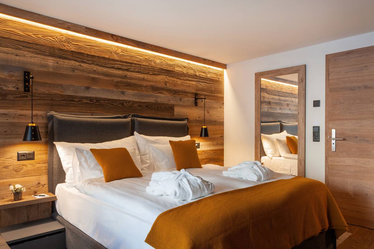 A modern hotel room with a wooden headboard, a large mirror, and a neatly made bed with white linens and orange accents.