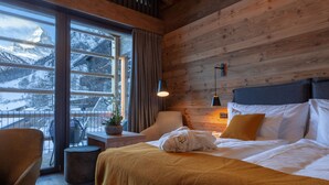 Ein gemütliches Zimmer mit Bett, Sessel, kleinem Tisch und Fenster mit Blick auf schneebedeckte Berge.