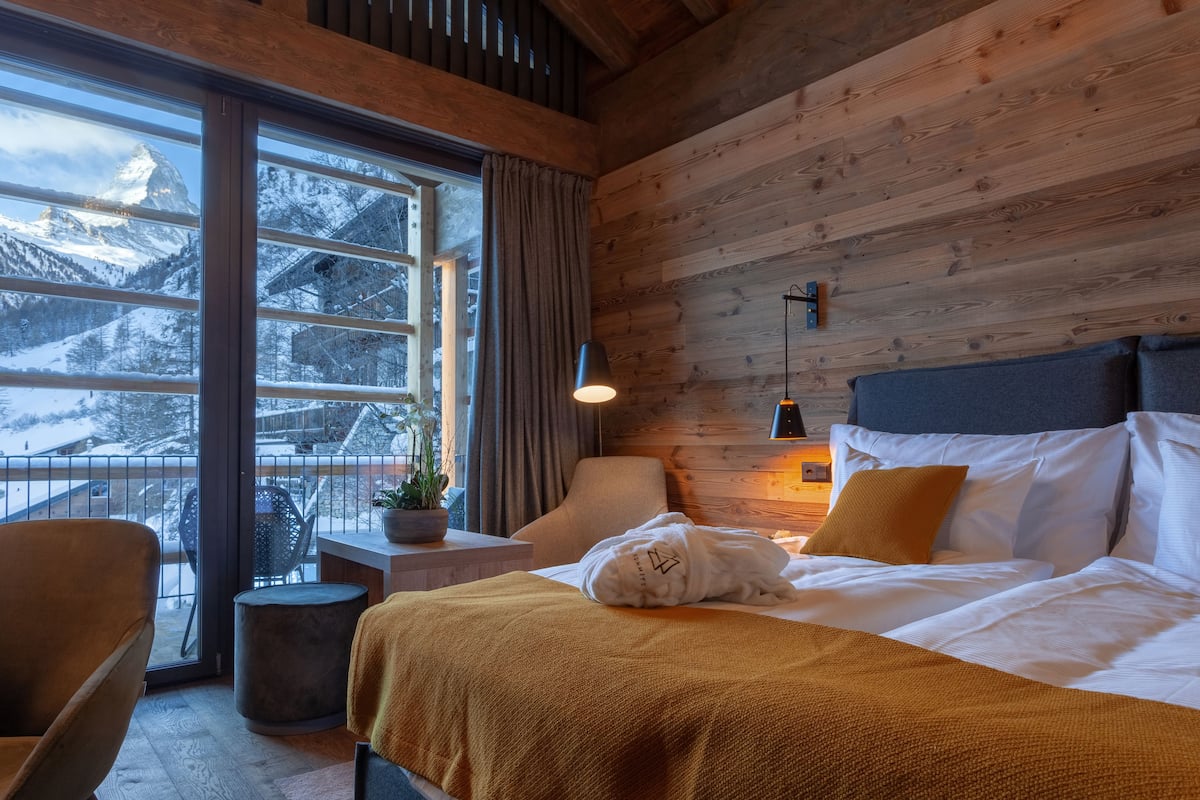 A cozy room with a bed, a chair, a small table, and a window with a view of snow-covered mountains.
