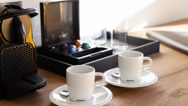 Two white Nespresso cups on saucers, a black coffee machine, and a box of colorful capsules on a wooden table. - 22 Summits Boutique Hotel (Zermatt)