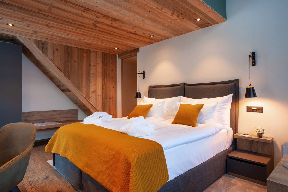 A modern bedroom with a wooden ceiling, a large bed with white linens and orange accents, a nightstand with a lamp, and a chair.