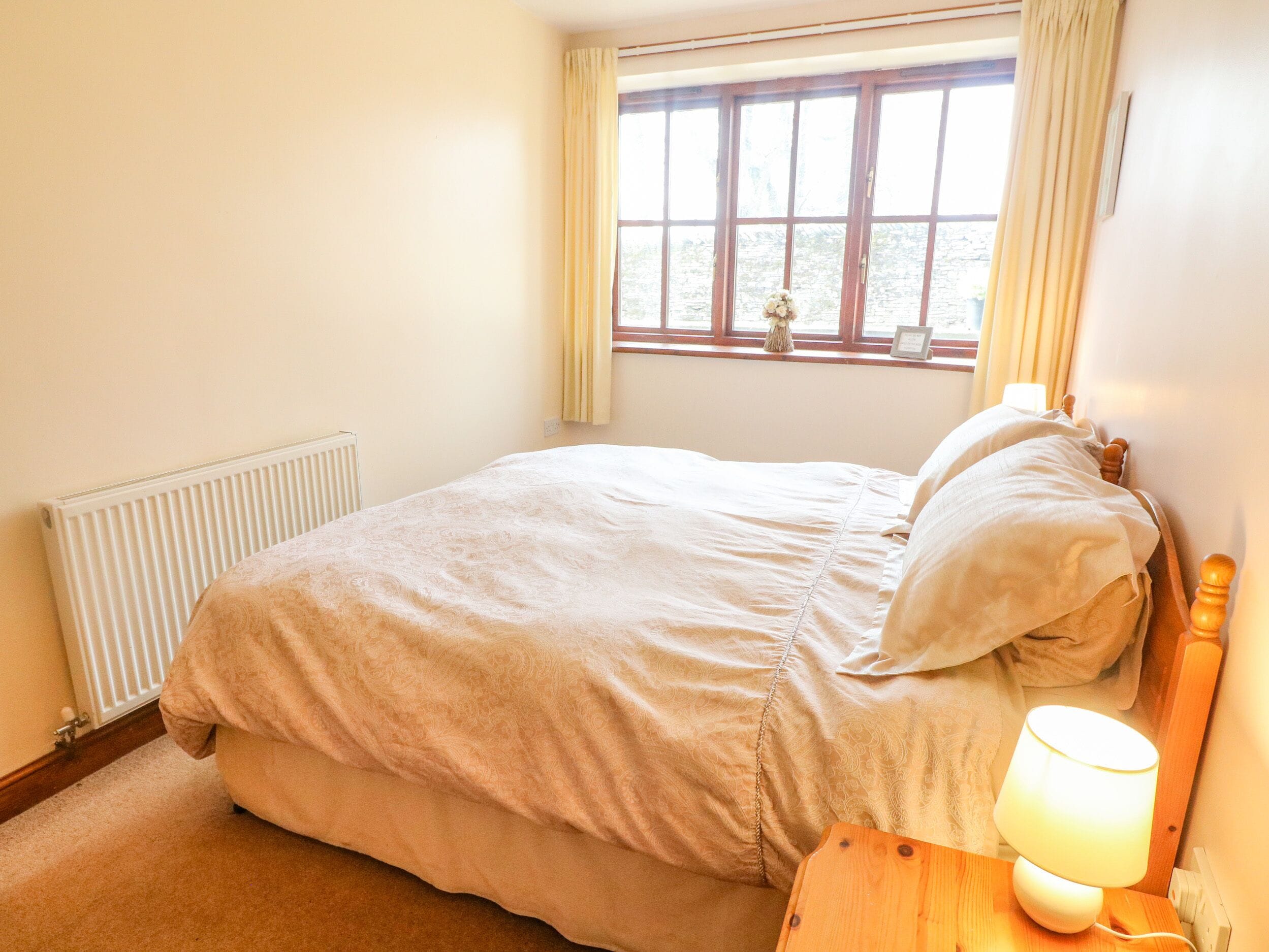A bedroom with a bed, a nightstand, a lamp, a window with curtains, and a radiator.