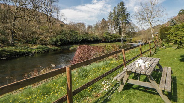 A riverside picnic area with a wooden fence, a bench, and a table with cups.