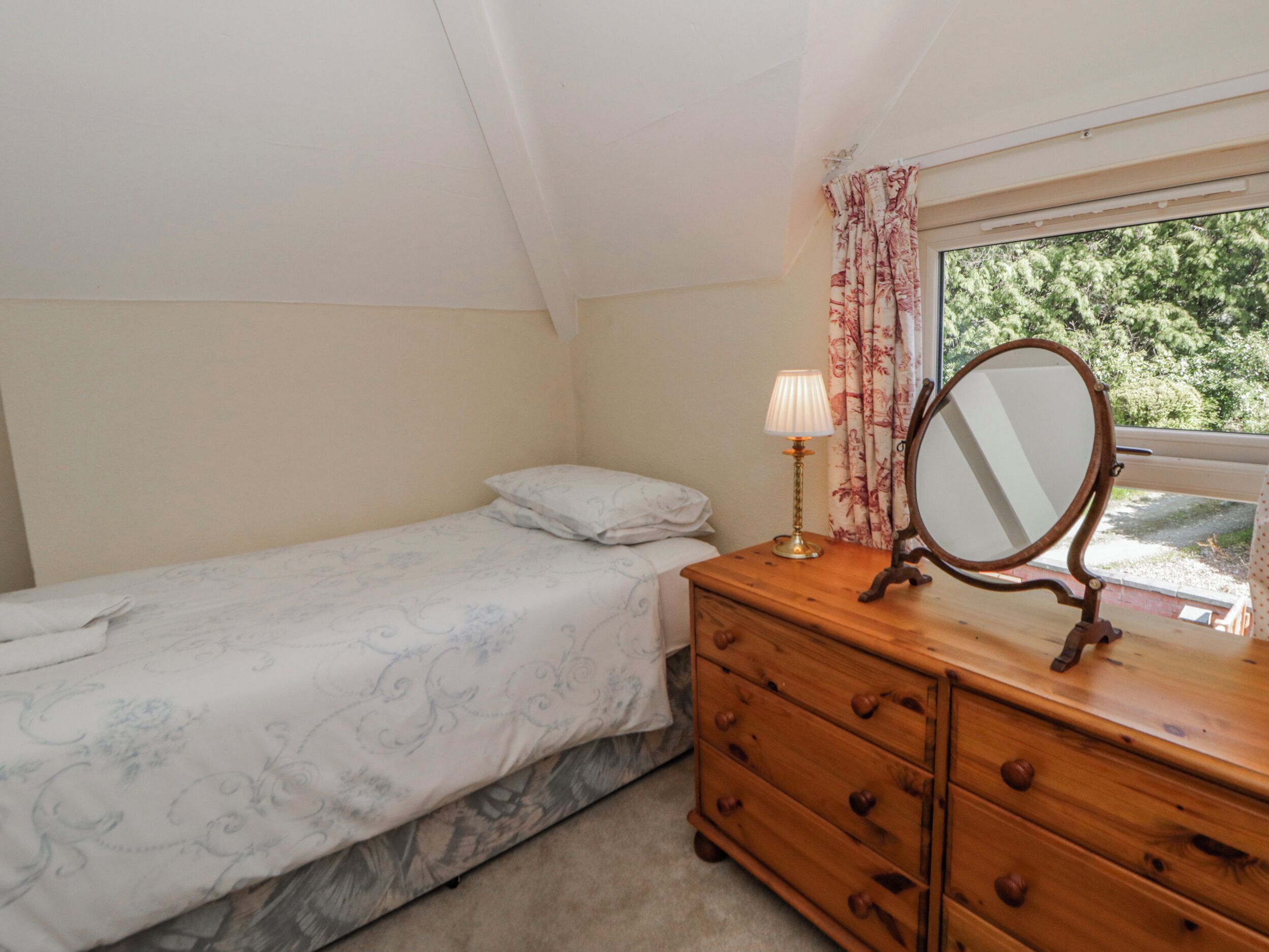 Une chambre avec un lit simple, une commode en bois avec des tiroirs, une lampe de chevet, un miroir et une fenêtre avec des rideaux.