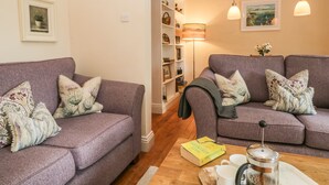 Un salon chaleureux avec deux canapés violets, une table basse sur laquelle reposent un livre et une cafetière, et une bibliothèque remplie de divers objets.
