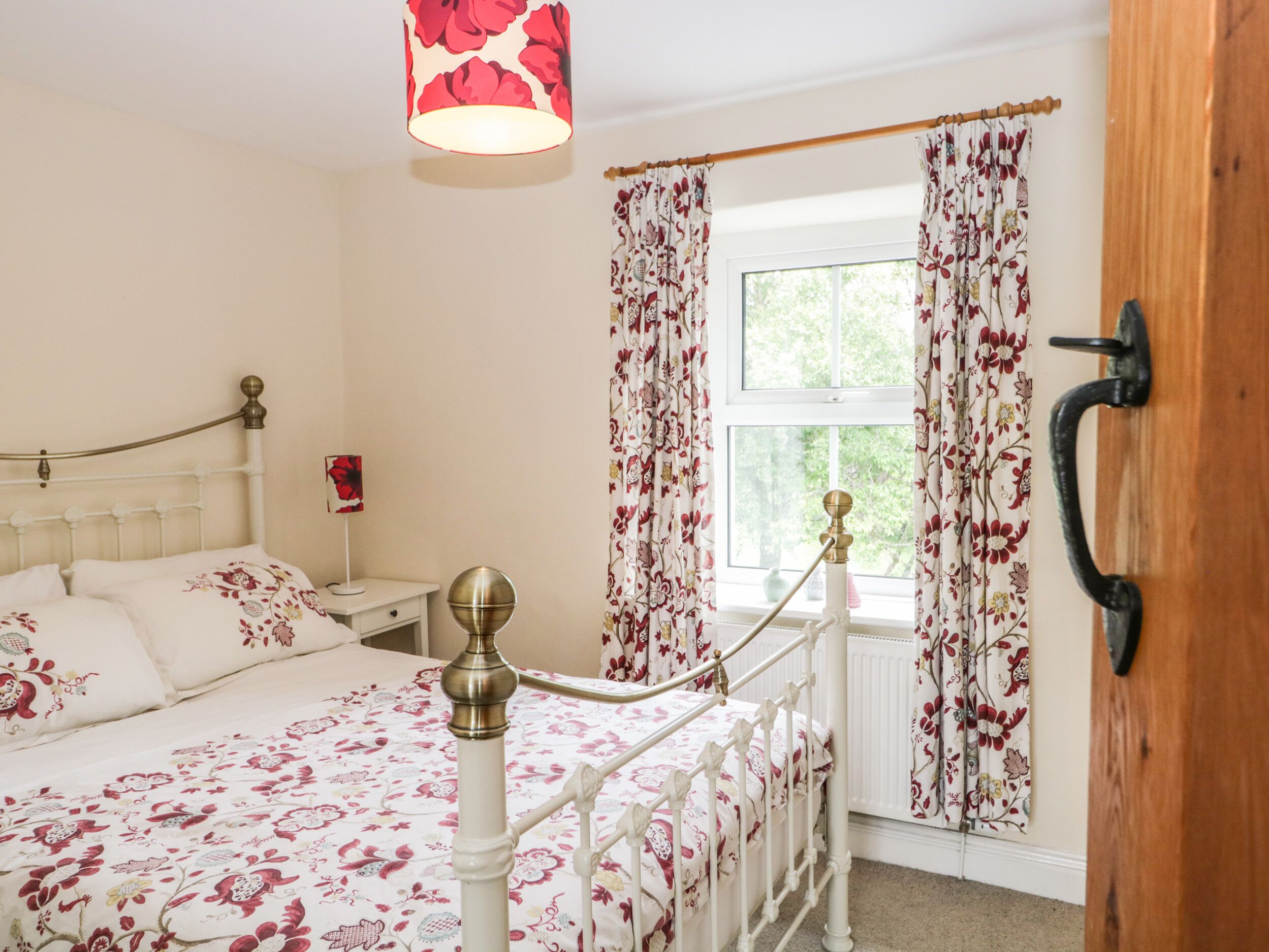 A bedroom with a floral bedspread, a bedside lamp, a window with curtains, and a wooden door with a handle.