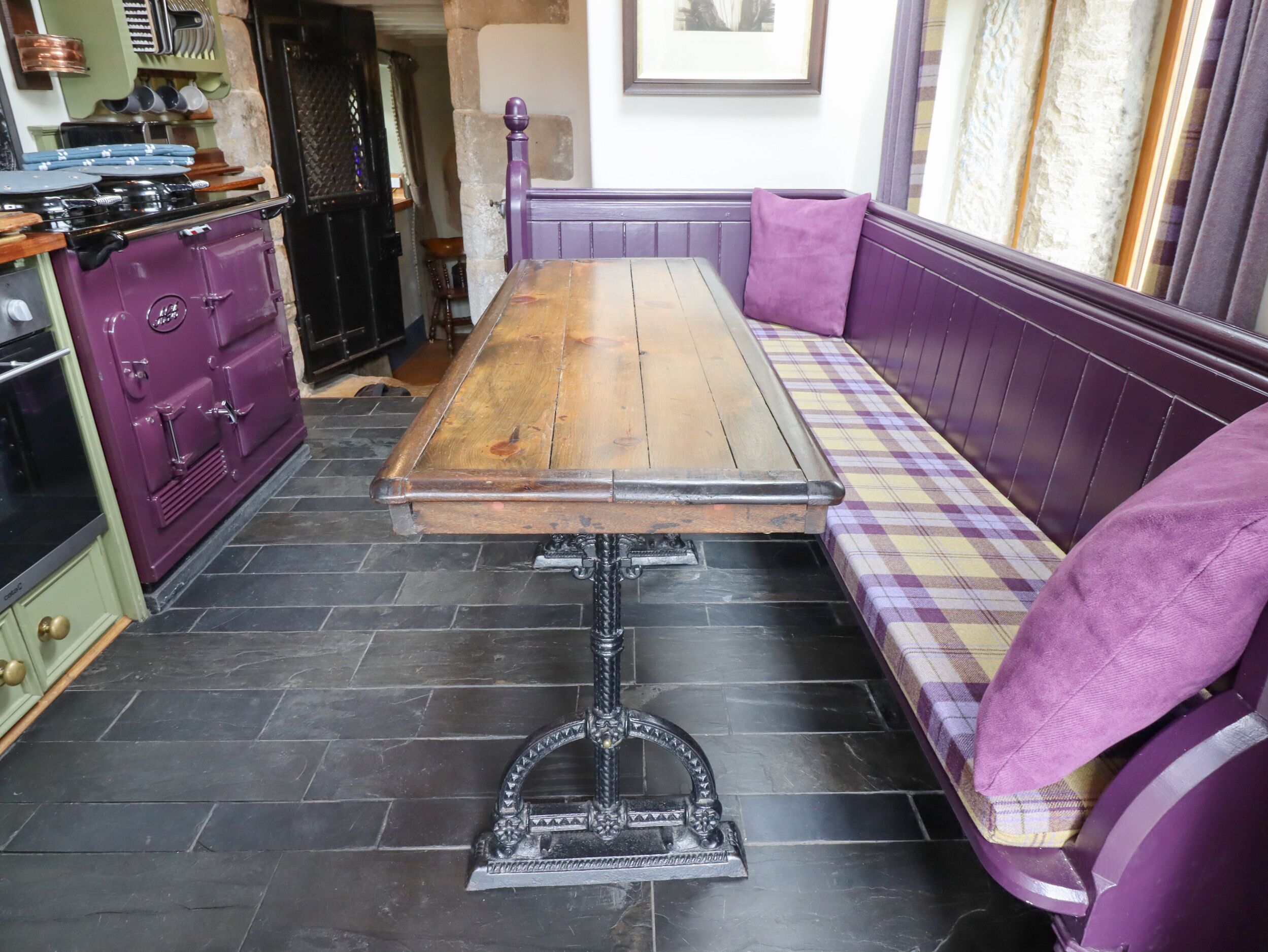 A kitchen with a wooden table and bench, a purple stove, and a checkered cushion.