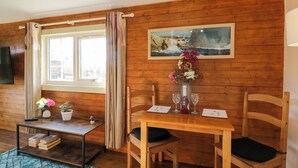 A wooden-paneled room with a table set for two, a window with curtains, a small table with a vase of flowers, and a painting on the wall.