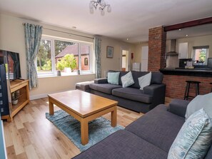 A living room with a sofa, a coffee table, a TV, and a kitchen area visible through an open door.