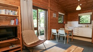 A cozy cabin interior with a wooden bookshelf, a television, a comfortable chair, a dining table, and a kitchen area. - Grasmere (Ulverston)